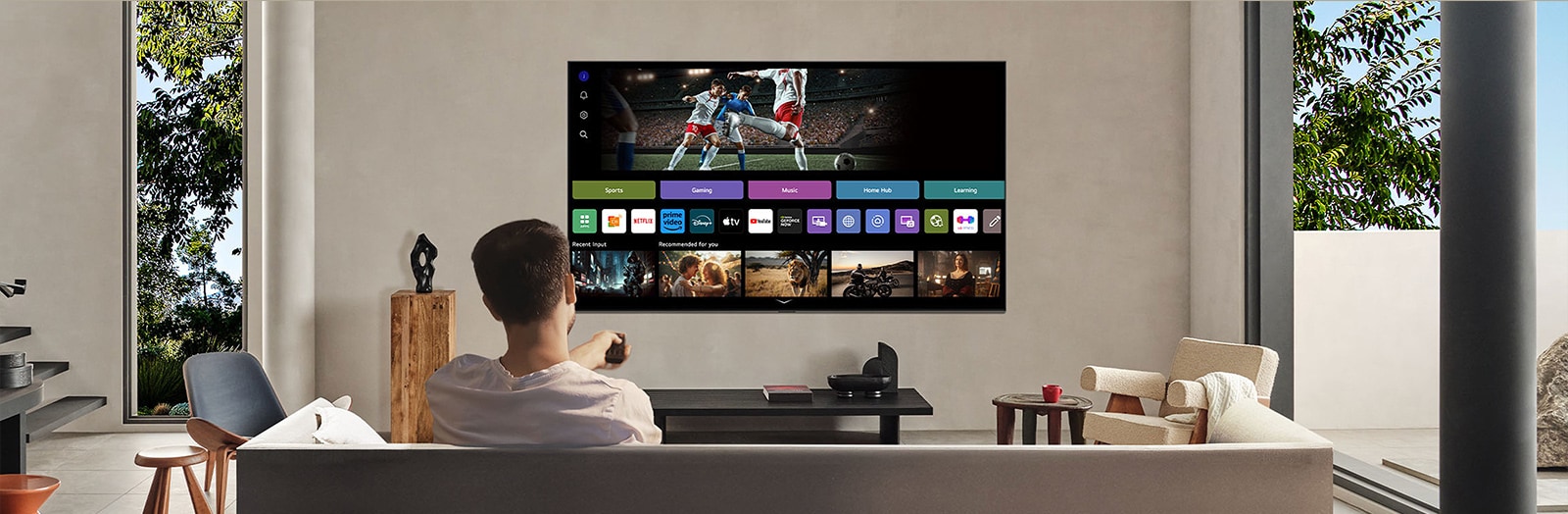 شخص على الأريكة يحمل جهاز تحكم عن بُعد. تلفزيون LG AI TV على الحائط مع LG webOS على الشاشة.