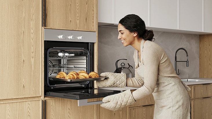 Woman placing a tray of baked pastries into a built-in oven.