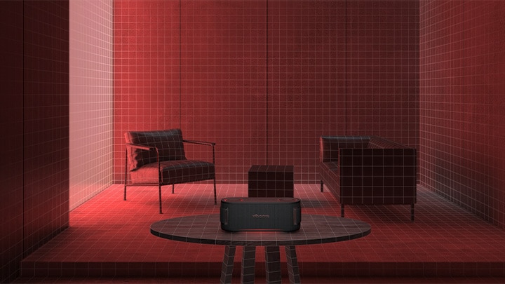 In a room filled with a red lighting a black sofa, a black ottoman  and a black armchair are placed behiind. In the fornt, xboom Bounce is placed on the top of black round table.