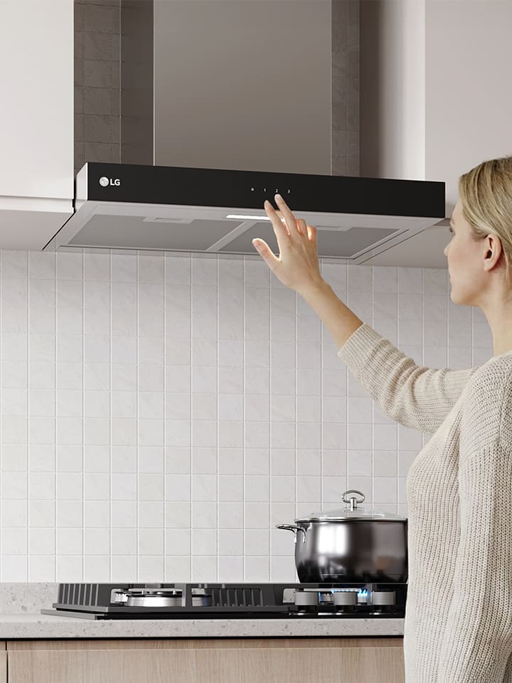 A woman adjusting the touch controls of a built-in range hood above a cooktop in a modern kitchen with a white tile wall.