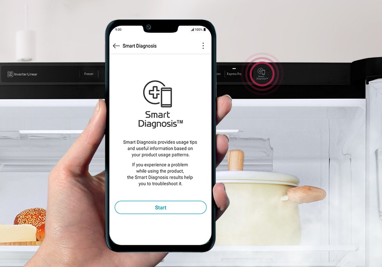 A hand holds a phone facing the front with a refrigerator in the background. 