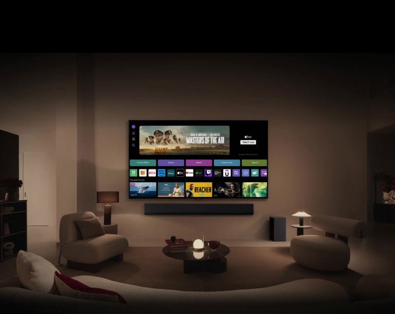 A close-up of an LG TV screen showing the buttons Home Office, Game, and Music over a banner for Masters of the Air zooms out to show the TV mounted on a wall in a living room. The following logos are displayed on the TV screen in the image: LG Channels, Netflix, Prime Video, Disney TV, Apple TV, YouTube, Spotify, Twitch, GeForce Now and Udemy.