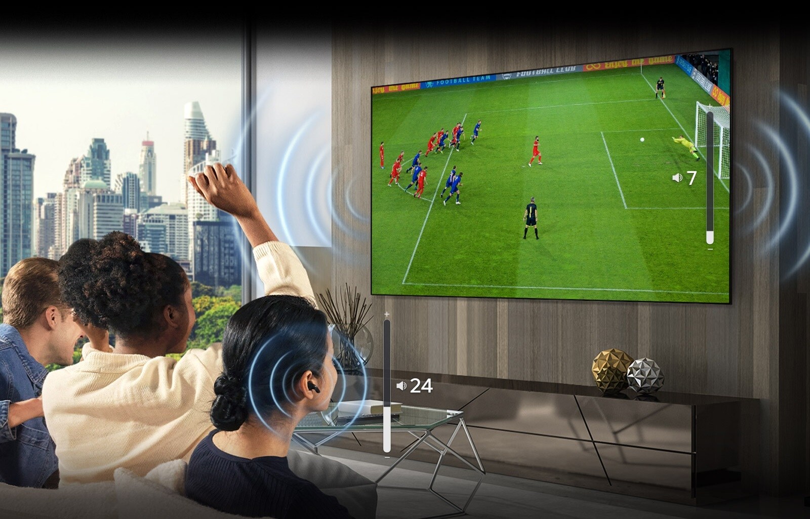A group of people are sitting on a sofa watching a soccer game on TV. The woman on the far right is wearing earbuds and using them with a different volume than the TV, indicating that she is using both at the same time.