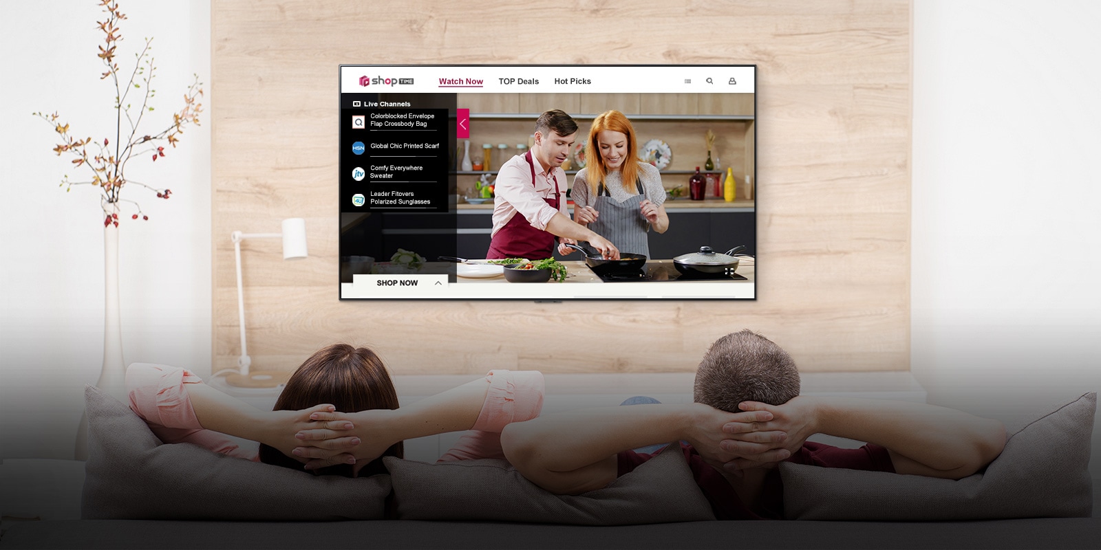 Couple sitting on a couch watching a TV displaying a man and a woman cooking in a kitchen and program information to shop.