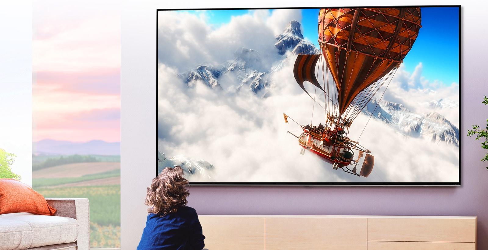 A child sitting in front of a large wall-mounted TV in a light room. The screen shows a hot air balloon flying through the clouds in front of a mountain.