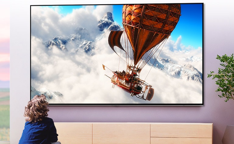 A child sitting in front of a large wall-mounted TV in a light room. The screen shows a hot air balloon flying through the clouds in front of a mountain.
