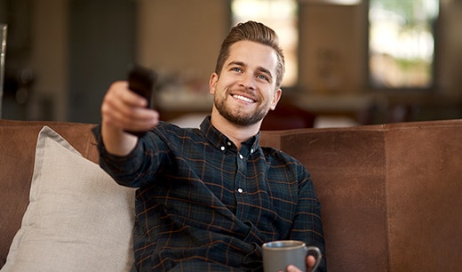 A man sitting on the sofa, holding a remote control with bright smile. The other hand is holding a mug.