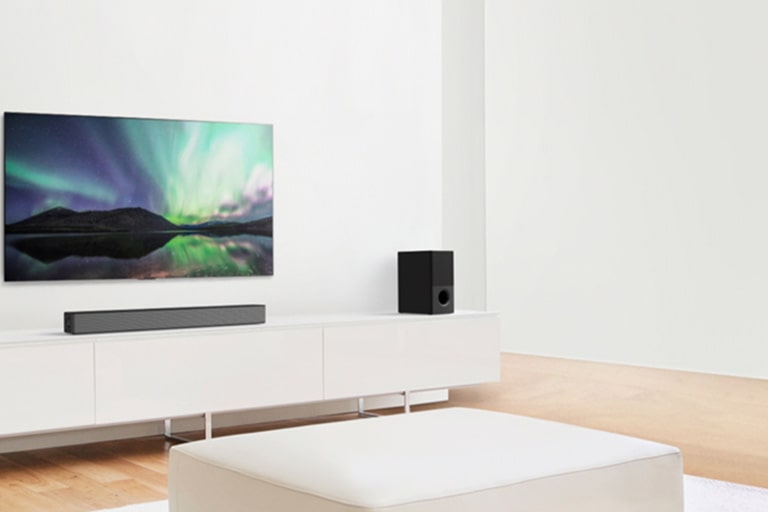 TV and sound bar in white living room with white sofa in the center. Speakers sit on both ends of the sofa.