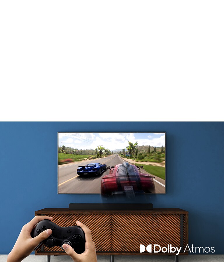  LG TV is on the wall, showing a racing game. LG Sound Bar is place on the brown shelf, right below LG TV. A man is holding a joy stick.