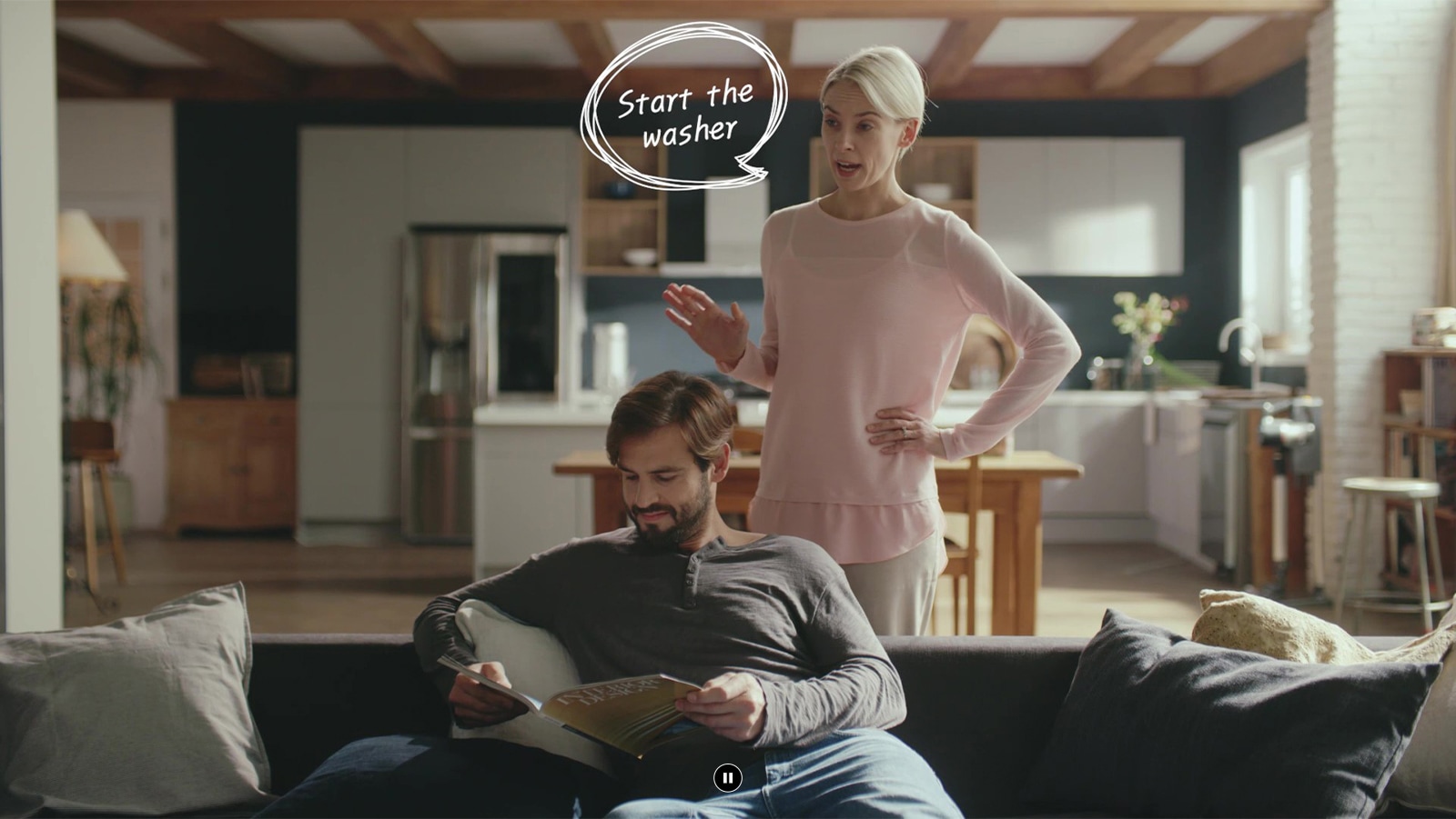 A woman is standing and talking to a man sitting on a sofa reading a book in the living room