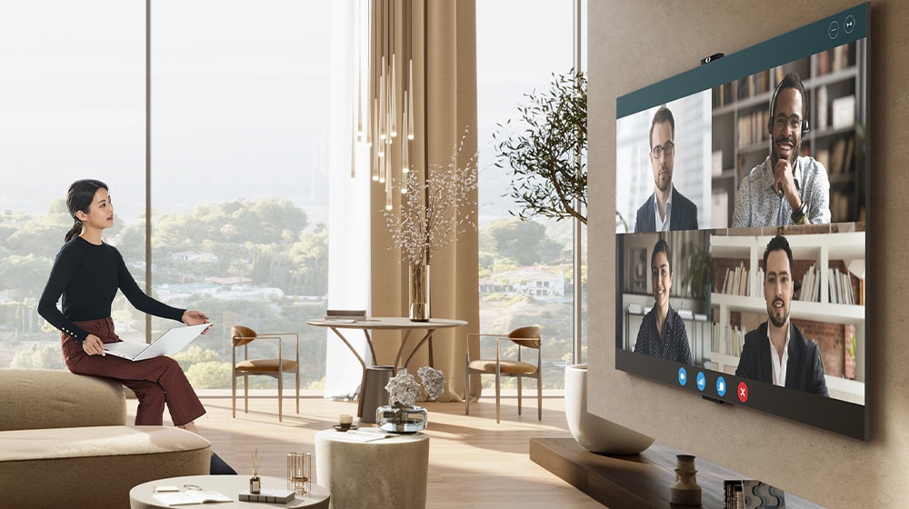 Moteris sėdi ant sofos porankio, laiko nešiojamąjį kompiuterį ir žiūri televizorių. Dideliame televizoriaus ekrane rodomi keturi žmonės ir vaizdo konferencija.