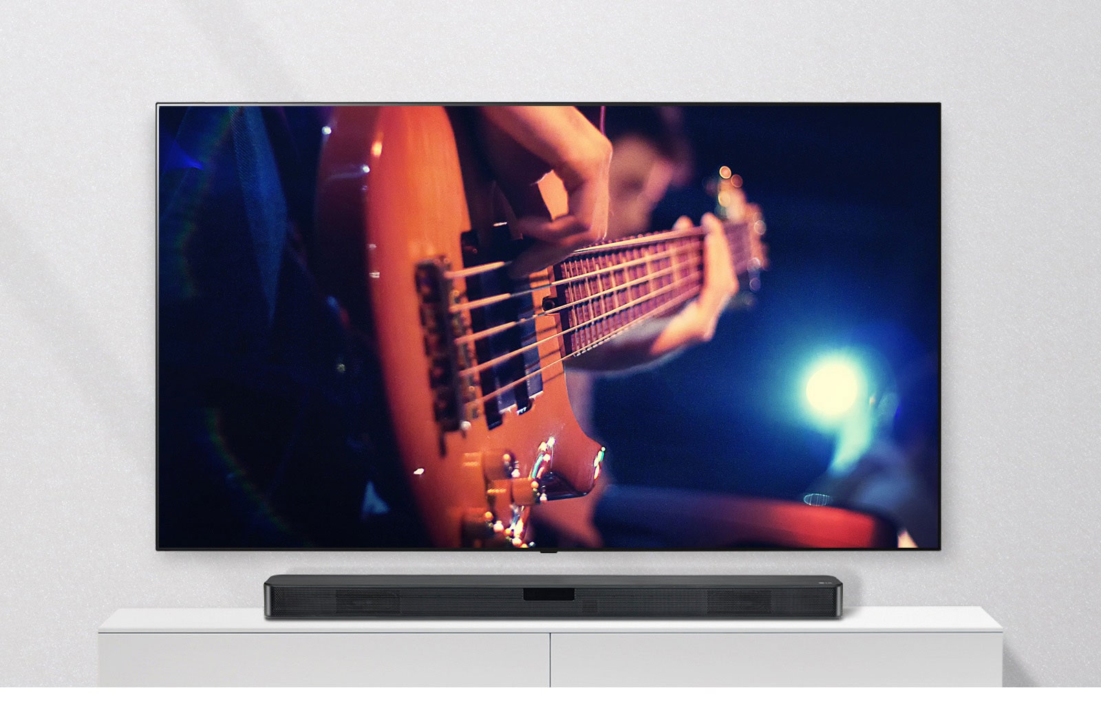 TV įrengtas ant sienos, o horizontalusis garsiakalbis padėtas ant baltos lentynos. Per TV rodomas gitara grojantis vyras.