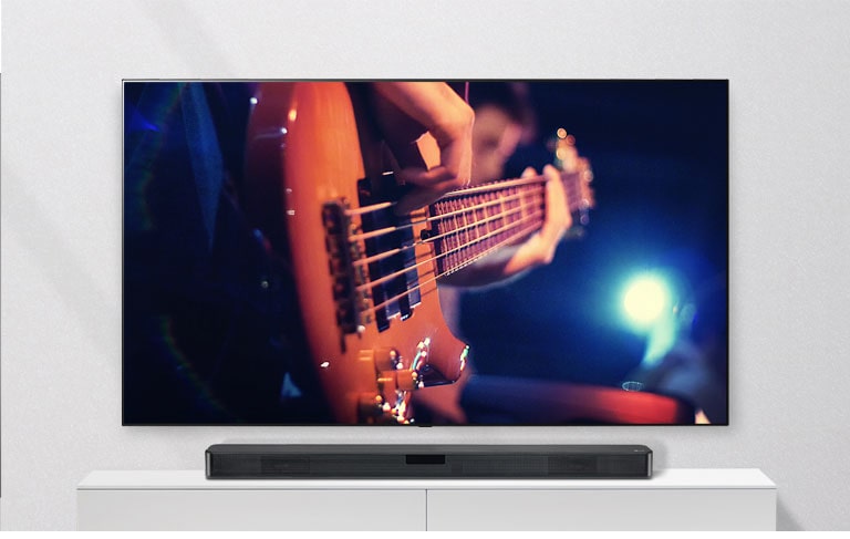 TV įrengtas ant sienos, o horizontalusis garsiakalbis padėtas ant baltos lentynos. Per TV rodomas gitara grojantis vyras.