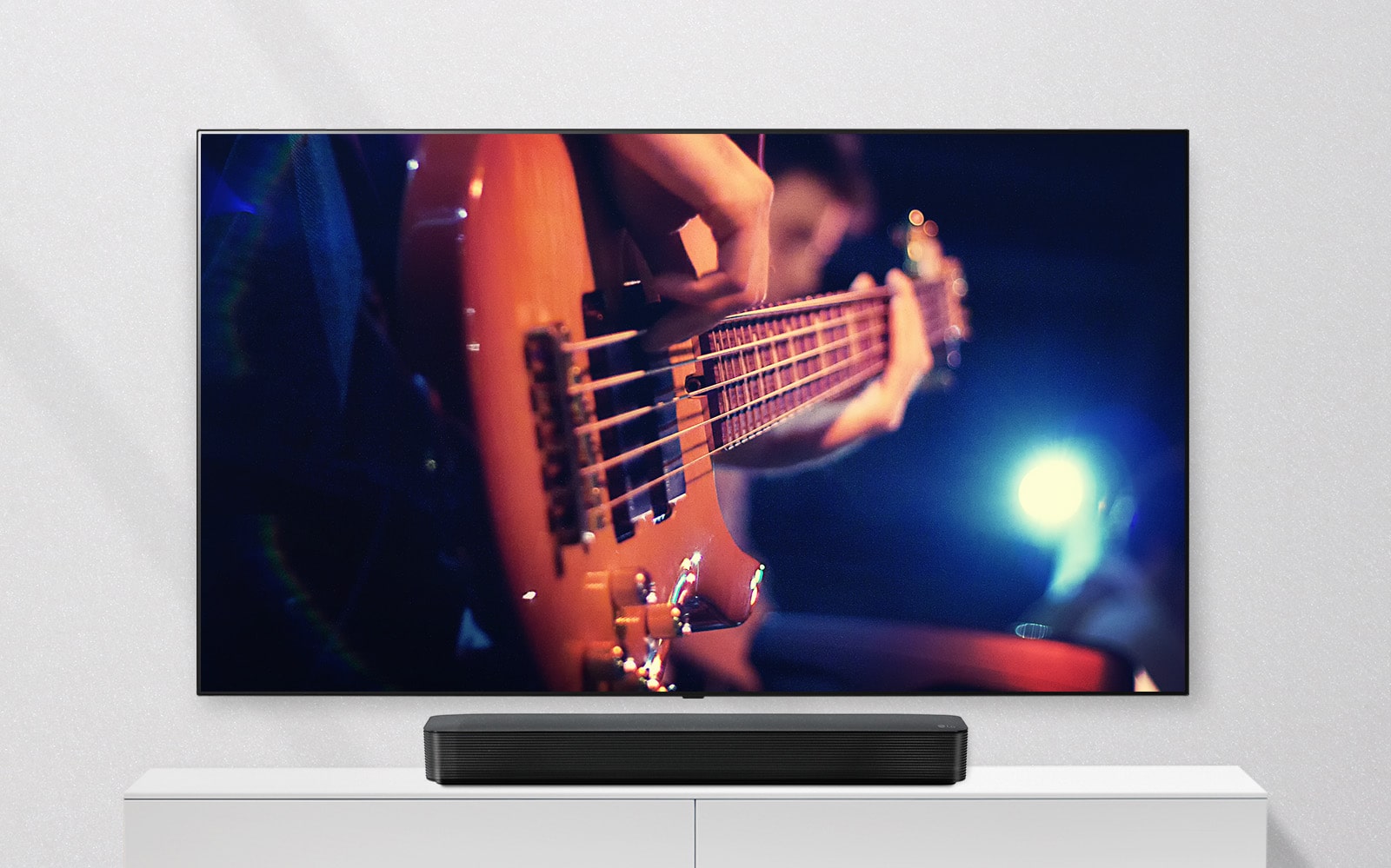 TV pritvirtintas prie sienos, o soundbar stovi ant baltos lentynos. TV rodomas gitara grojantis vyras.