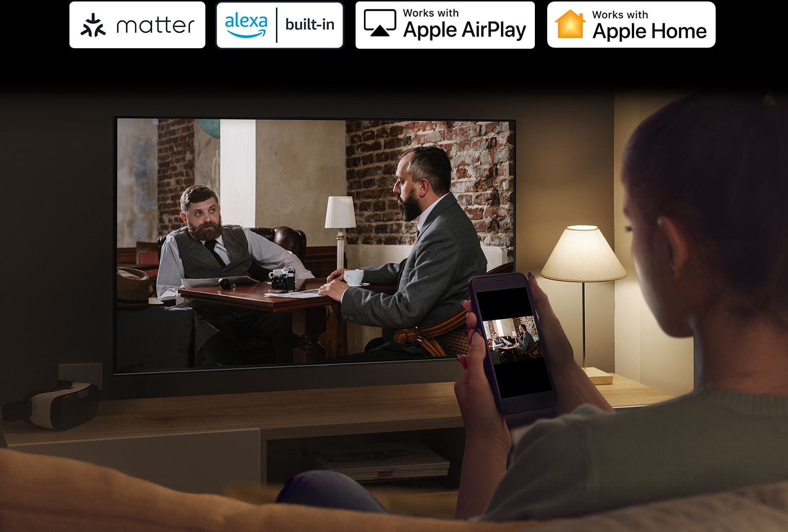 Tamsiame kambaryje matyti moteris, kuri žiūri vaizdo įrašą iš savo telefono televizoriuje, naudodama ekrano atvaizdavimo funkciją.