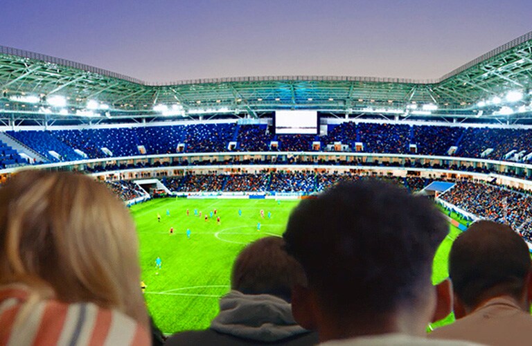 Žmonės dideliame TV žiūri futbolo rungtynes, fone matomas visas stadionas