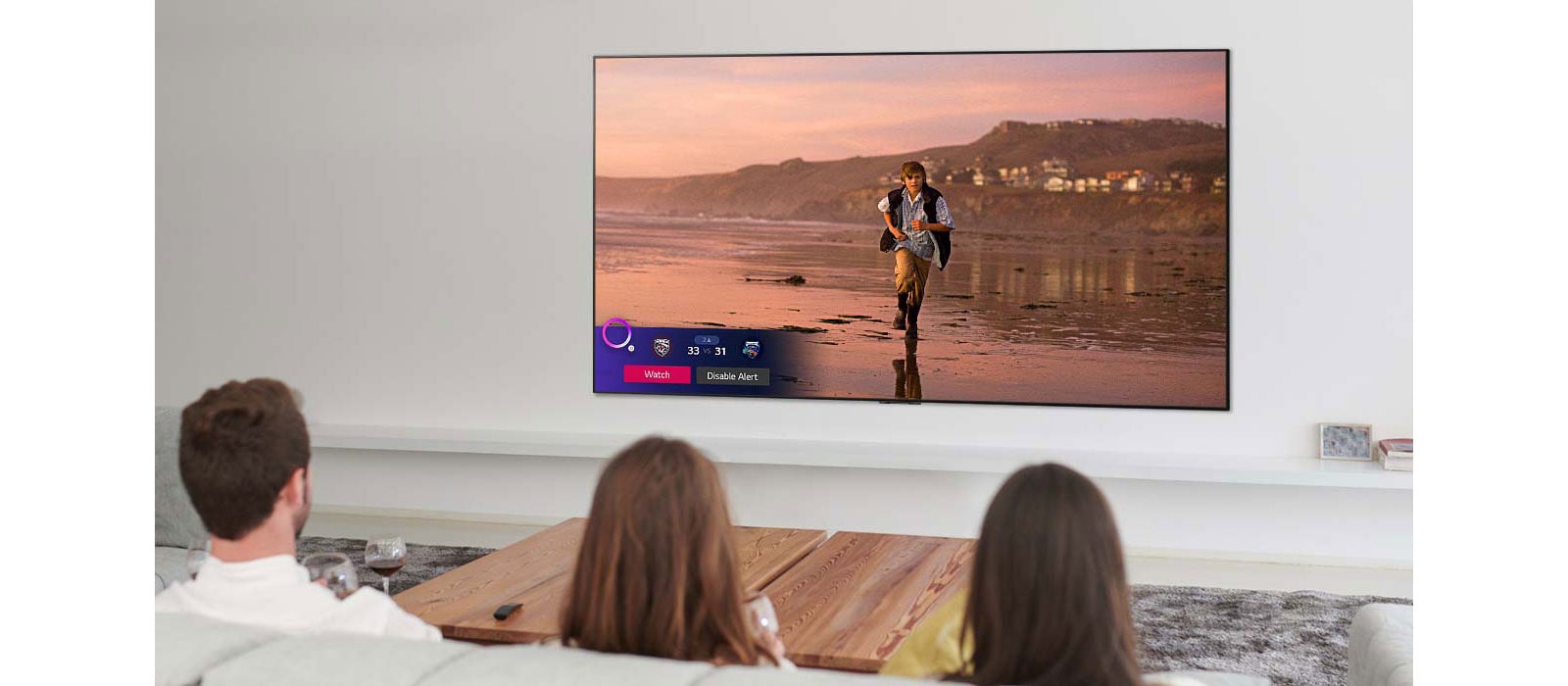 Trys žmonės žiūri TV ekrane rodomą scena iš magiškosios fantastikos filmo, taip pat rodomas su sportu susijęs įspėjimas