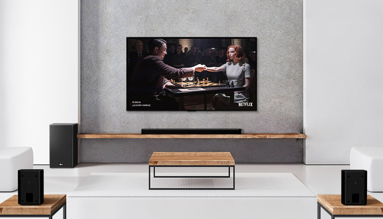 Rinkinys, kurį sudaro 2 užpakaliniai garsiakalbiai, žemadažnis garsiakalbis, horizontalusis garsiakalbis ir televizorius baltoje svetainėje. Televizoriaus ekrane moteris ir vyras žaidžia šachmatais.