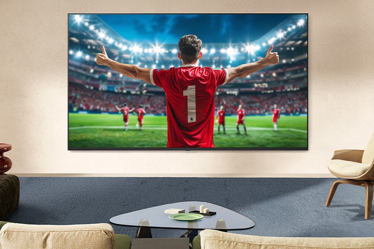 LG NANO 4K UHD AI NU80 Ultra Big TV ekranas pritvirtintas prie sienos kambaryje rodo futbolininką, švenčiantį pergalę stadiono centre, apšviestą ryškių arenos žibintų; pabrėžiamos sodrios spalvos, raiška ir įtraukiantis mastelis.