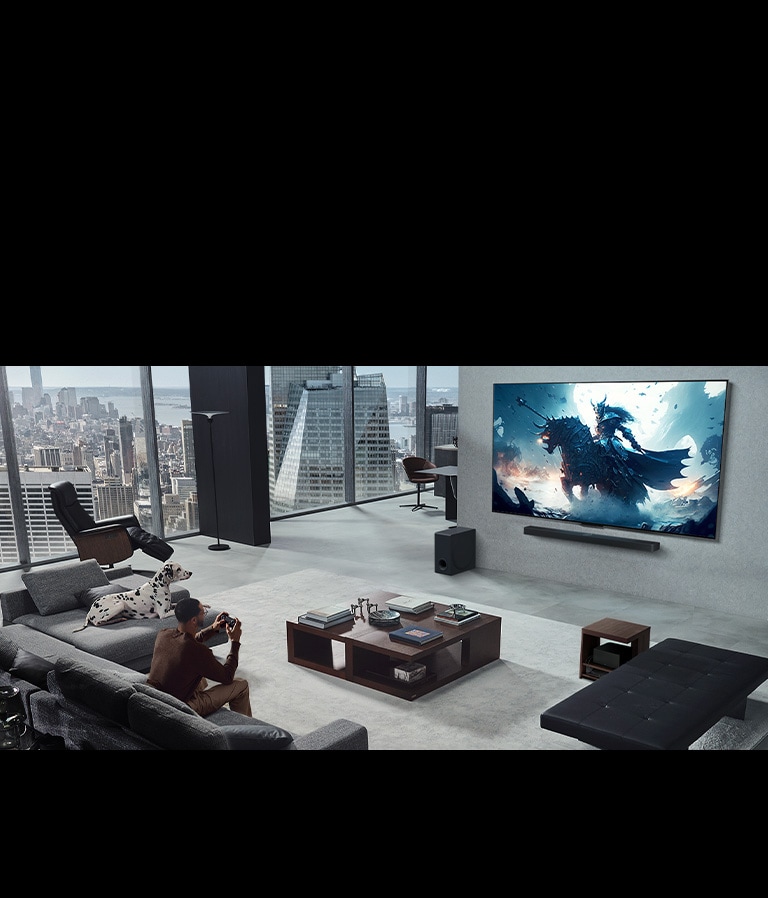 Kambarys, kuriame žmogus žaidžia vaizdo žaidimą per ant sienos sumontuotą „LG True Wireless TV“ ir klausosi per „LG Soundbar“ garso sistemą. Rankoje jis laiko valdiklį, tačiau žaidimų konsolės iš pirmo žvilgsnio nematyti. Taip pat nematyti jokių laidų. „Zero Connect Box“ paslėptas ir vos matomas po staliuku. 