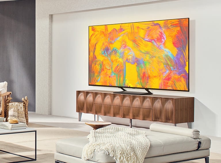 LG QNED TV stovi ant spintelės jaukioje gyvenamojoje erdvėje. LG QNED TV rodomas abstraktus oranžinės, geltonos, violetinės ir rožinės spalvų fonas.