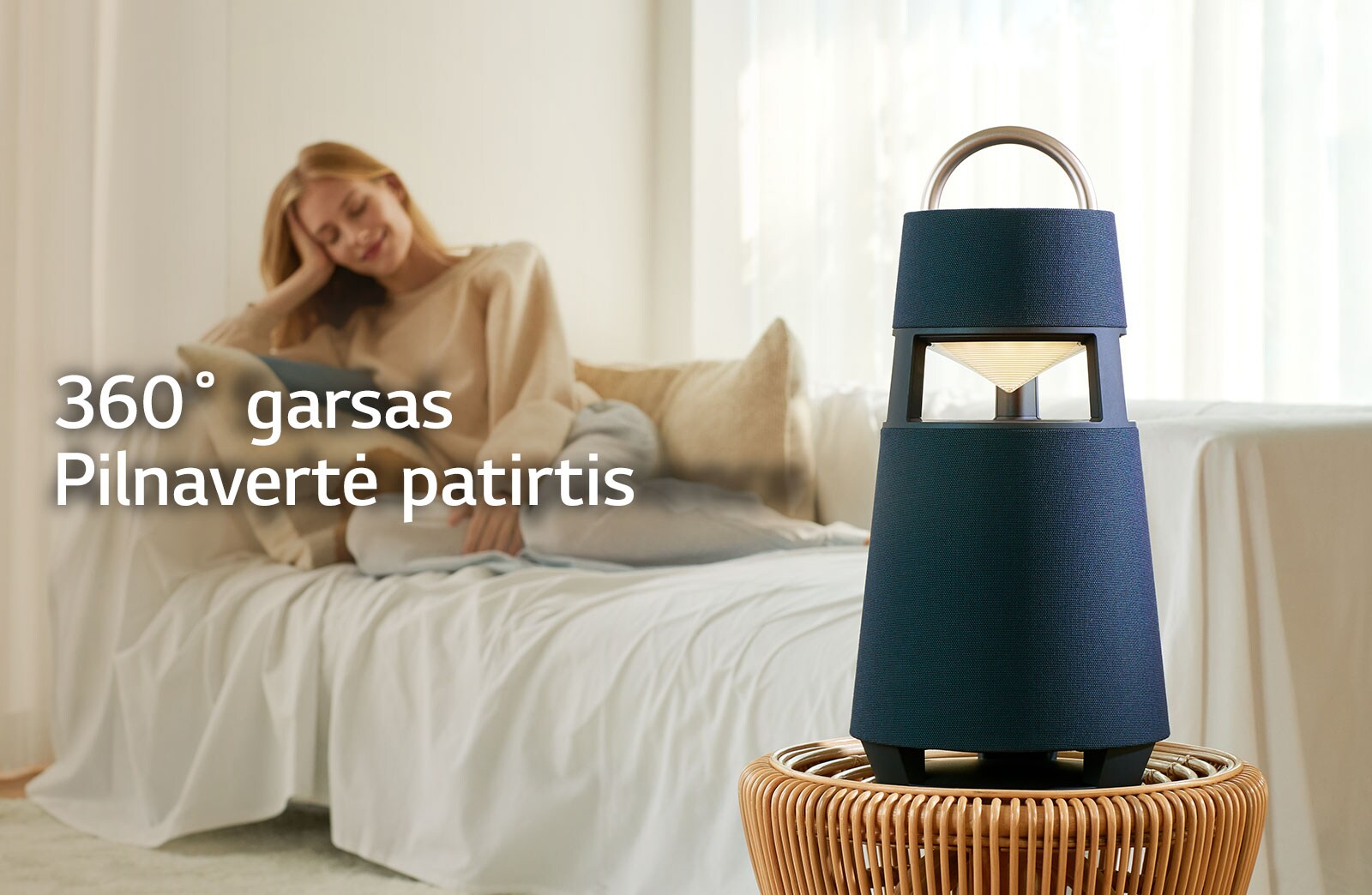 Paveikslėlyje matoma ant sofos sėdinti moteris, besiklausanti muzikos, sklindančios iš prieš ją stovinčio XBOOM 360 garsiakalbio.