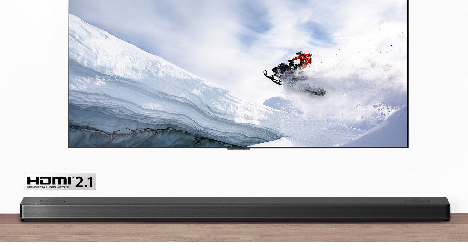 Televizoriaus ir horizontaliojo garsiakalbio vaizdas iš priekio. Per televizorių rodomas snieguotu kalnu ant sniegaeigio važiuojantis žmogus. HDMI 2.1 logotipas po televizoriumi.