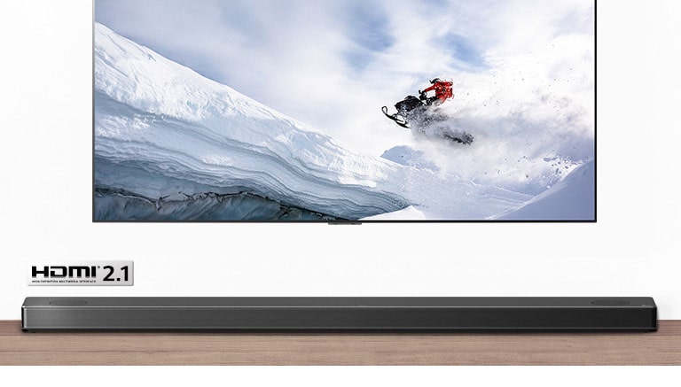 Televizoriaus ir horizontaliojo garsiakalbio vaizdas iš priekio. Per televizorių rodomas snieguotu kalnu ant sniegaeigio važiuojantis žmogus. HDMI 2.1 logotipas po televizoriumi.