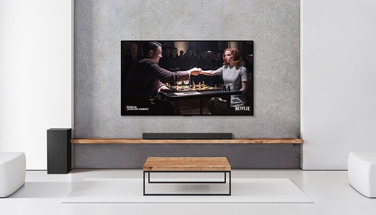 Žemadažnis garsiakalbis, horizontalusis garsiakalbis ir televizorius baltoje svetainėje. Televizoriaus ekrane moteris ir vyras žaidžia šachmatais.