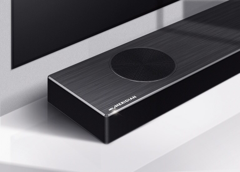 LG horizontaliojo garsiakalbio su „Meridian“ logotipu apatiniame kairiajame kampe kairės pusės vaizdas stambiu planu. Taip pat matoma televizoriaus apatinė kairioji pusė.