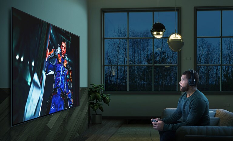 Vyras tamsiame kambaryje sėdi ant sofos priešais didelį TV ekraną ir žaidžia veiksmo žaidimą.