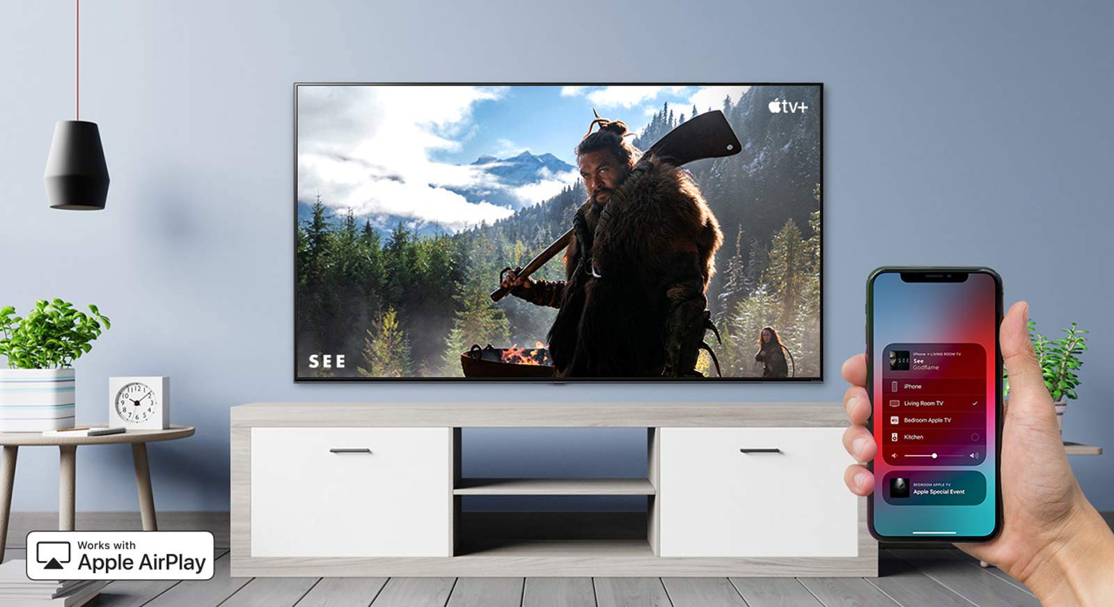 Televizorių „iPhone“ įrenginiu valdanti ranka stambiu planu ir televizorius, kuriame rodomas „Apple TV+“ turinys
