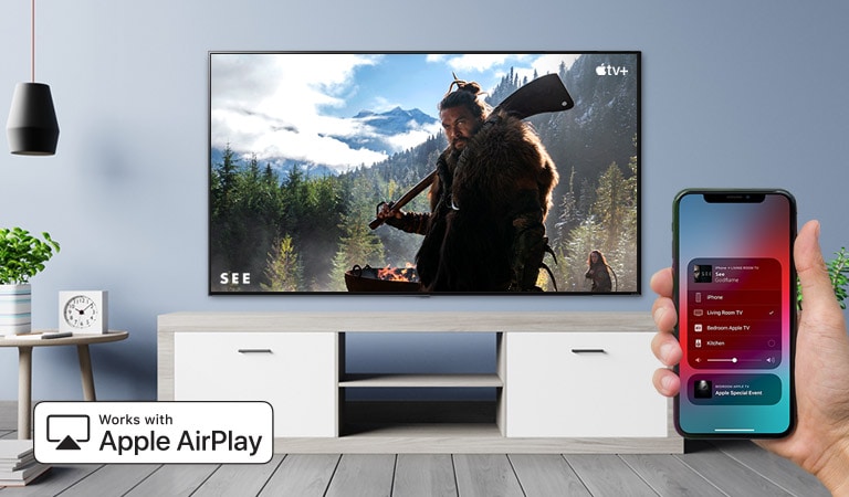 Televizorių „iPhone“ įrenginiu valdanti ranka stambiu planu ir televizorius, kuriame rodomas „Apple TV+“ turinys