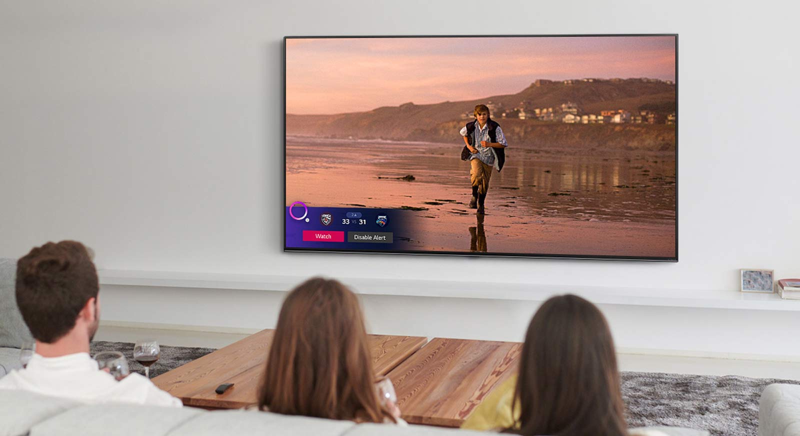 Trys žmonės žiūri TV ekrane rodomą scena iš magiškosios fantastikos filmo, taip pat rodomas su sportu susijęs įspėjimas