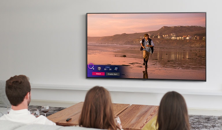 Trys žmonės žiūri TV ekrane rodomą scena iš magiškosios fantastikos filmo, taip pat rodomas su sportu susijęs įspėjimas