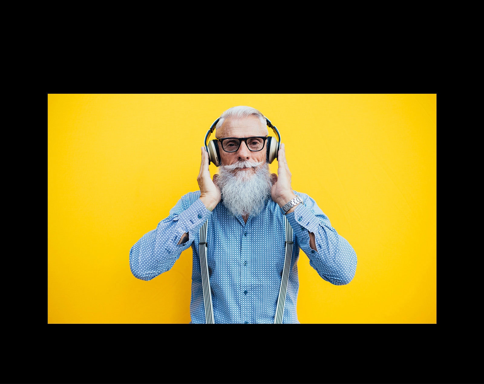 Priešais geltoną sieną sėdi senolis, dėvintis ausines.