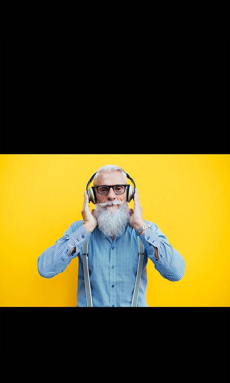 Priešais geltoną sieną sėdi senolis, dėvintis ausines.