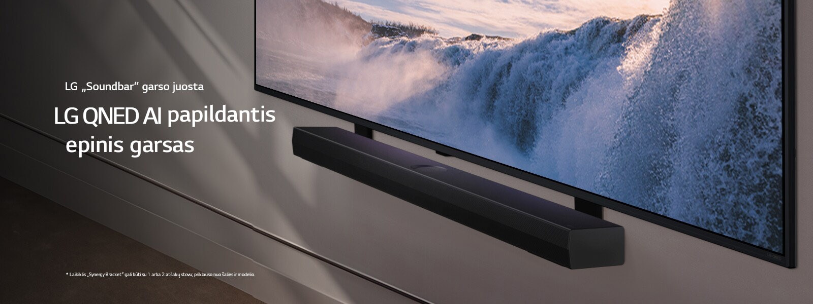 LG TV ir „Soundbar“ pritvirtinti prie sienos, vaizdas kampu. Televizoriuje iš arti rodomas didžiulis krioklys, o per sieną, televizorių ir garso juostą sklinda švelni saulės šviesa. Atsakomybės atsisakymo tekstas: Laikiklis „Synergy Bracket“ gali būti su 1 arba 2 atšakų stovu; priklauso nuo šalies ir modelio.