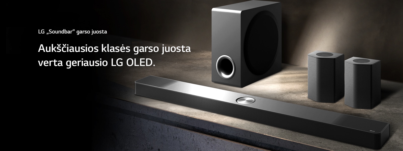 LG „Soundbar“ ir galiniai bei žemųjų dažnių garsiakalbiai išdėstyti kampu ant rudos medinės lentynos, juodoje tamsos apgaubtoje patalpoje, su vienintele šviesa, sklindančia virš garso sistemos.