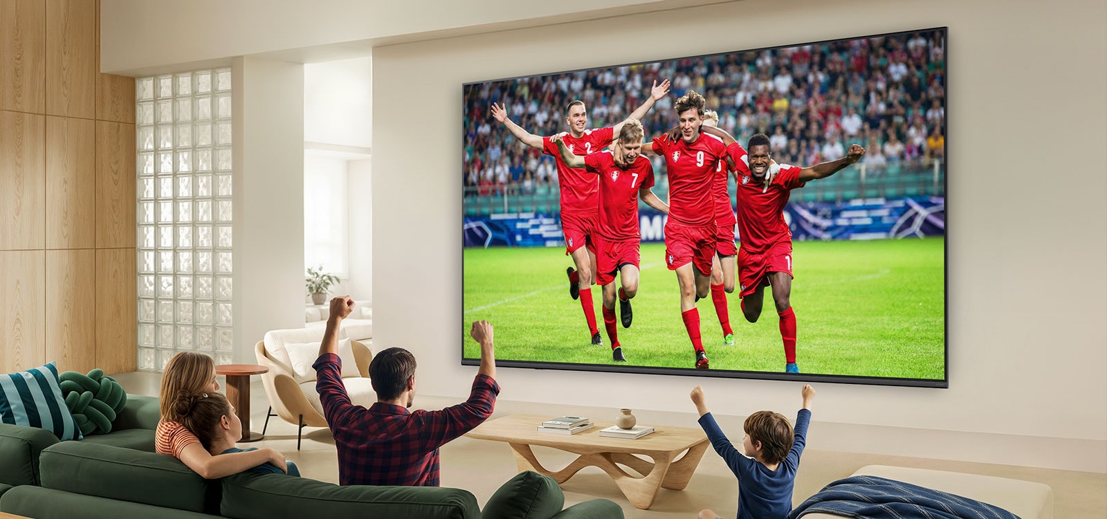 „LG QNED evo AI QNED80 Mini LED“ Ultra Big TV pritvirtintas prie sienos šviesiame gyvenamajame kambaryje su ant sofos kartu džiūgaujančia šeima; jo ekrane rodomas futbolo varžybų pergalės šventimas; ryškios spalvos ir rafinuota vaizdo kokybė plačiame ekrane.