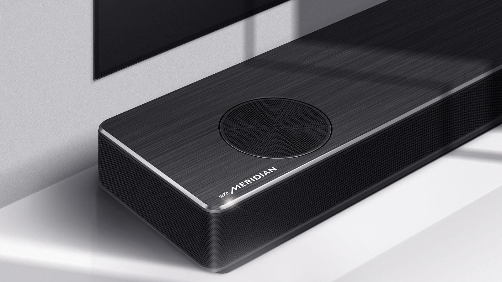 „LG Soundbar“ su „Meridian“ logotipu apatiniame kairiajame kampe kairės pusės vaizdas stambiu planu. Taip pat matoma televizoriaus apatinė kairioji pusė.