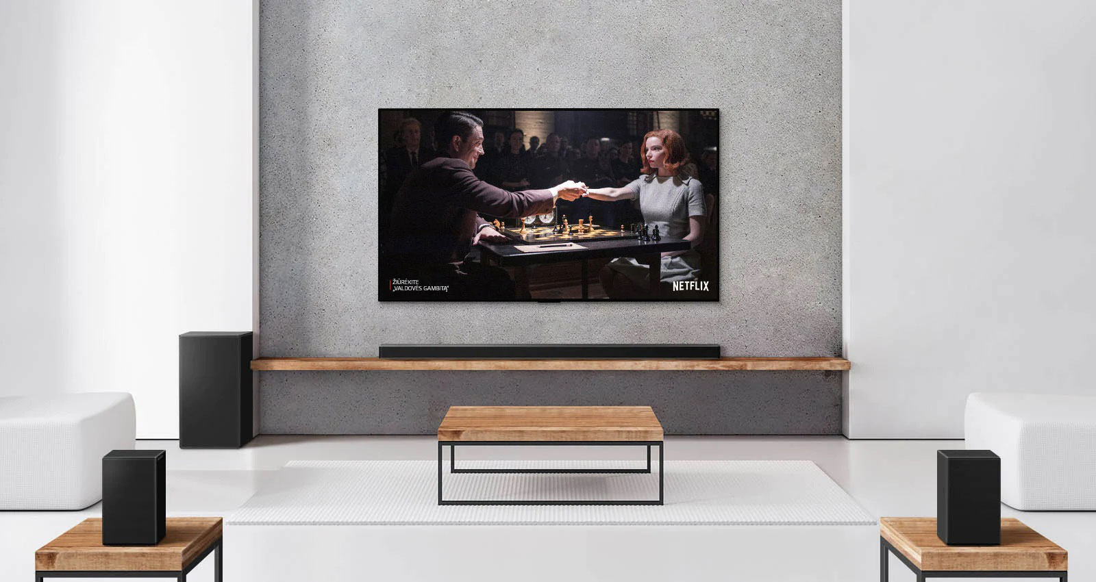 Rinkinys, kurį sudaro 2 užpakaliniai garsiakalbiai, žemadažnis garsiakalbis, „Soundbar“ ir televizorius baltoje svetainėje. Televizoriaus ekrane moteris ir vyras žaidžia šachmatais.