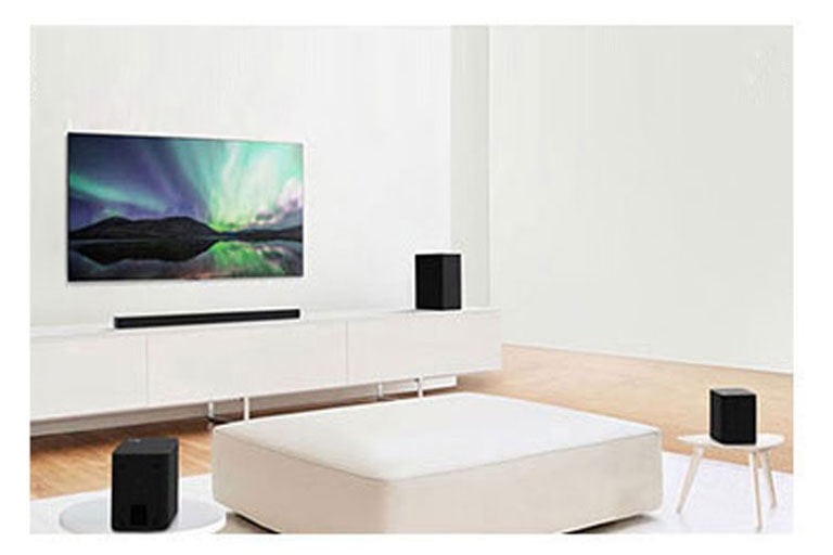 TV ir gulsčiasis garsiakalbis baltoje svetainėje, kurios centre yra sofa. Garsiakalbiai pastatyti abiejuose sofos galuose.