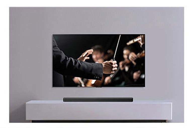 Televizorius įrengtas ant pilkos sienos, po juo ant pilkos lentynos padėtas „LG Soundbar“. Televizoriuje rodomas orkestrui diriguojantis dirigentas.