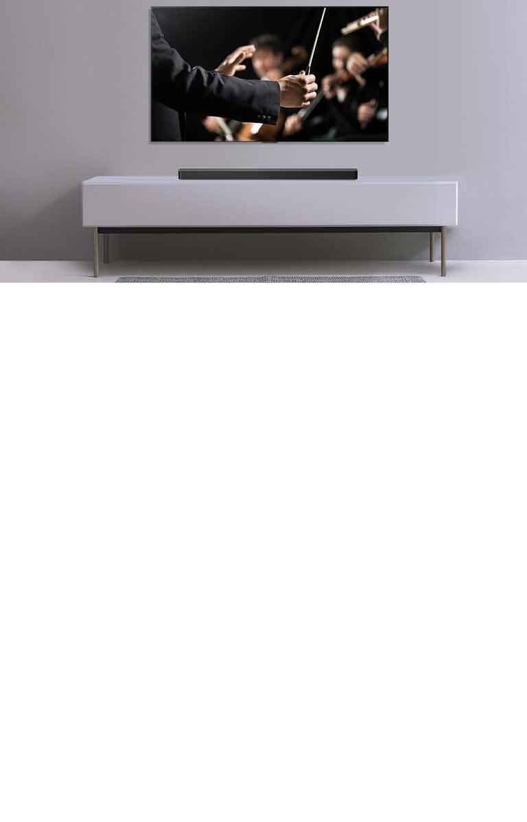 Televizorius įrengtas ant pilkos sienos, po juo ant pilkos lentynos padėtas „LG Soundbar“. Televizoriuje rodomas orkestrui diriguojantis dirigentas.