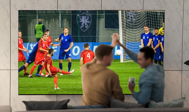 Žmonės svetainėje itin dideliame TV ekrane stebi sporto rungtynes