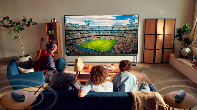 Žmonės svetainėje televizoriuje, prie kurio prijungti galiniai „Bluetooth“ garsiakalbiai, stebi sporto rungtynes