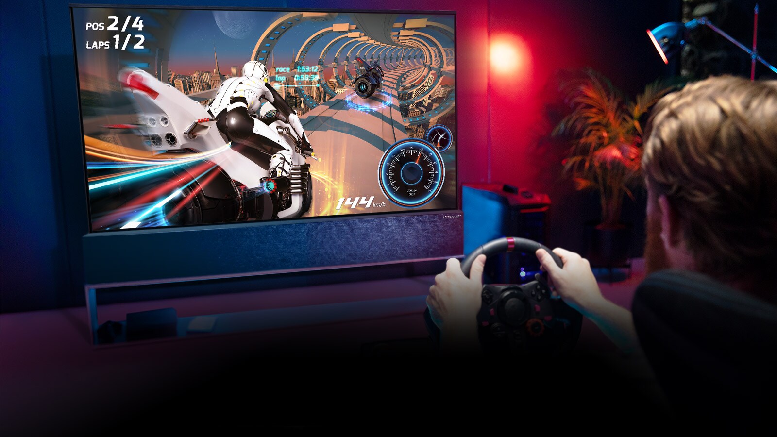 Vyras sėdi lenktynių žaidimų kėdėje, laiko lenktyninio automobilio vairą ir žaidžia lenktynių žaidimą TV ekrane