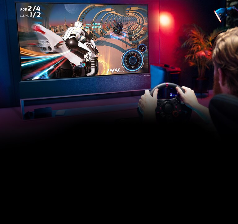 Vyras sėdi lenktynių žaidimų kėdėje, laiko lenktyninio automobilio vairą ir žaidžia lenktynių žaidimą TV ekrane