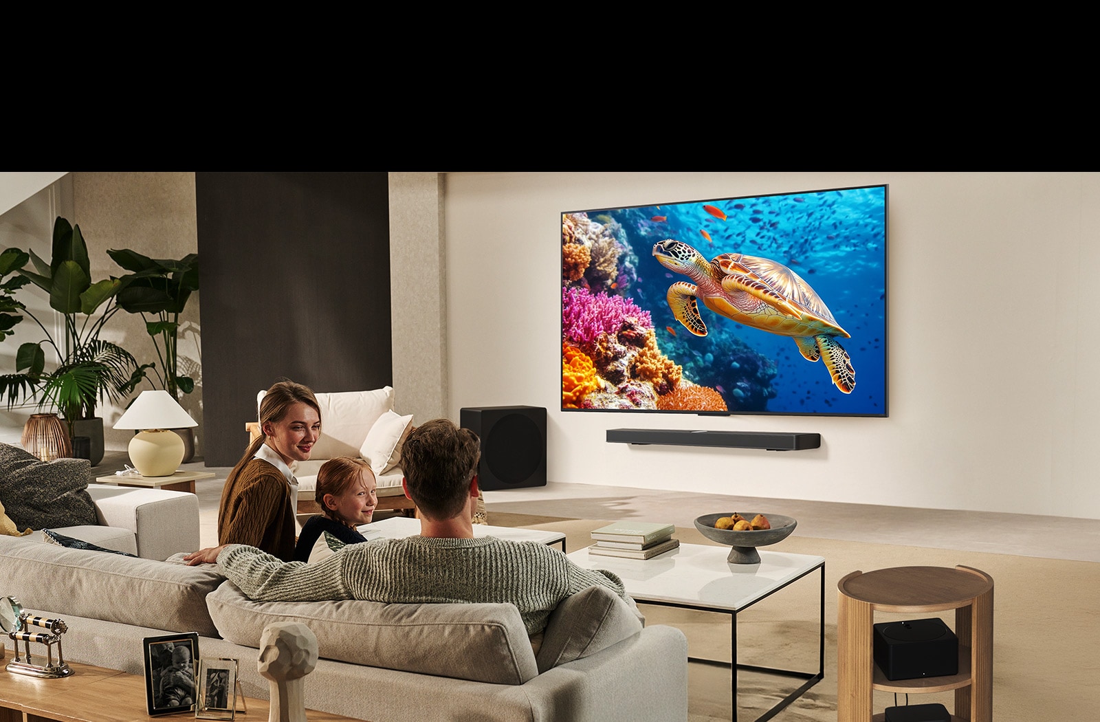 Šeima sėdi ant sofos priešais LG TV, sumontuotą ant sienos virš „LG Soundbar“. „Zero Connect Box“ yra kavos staliuko viduje.